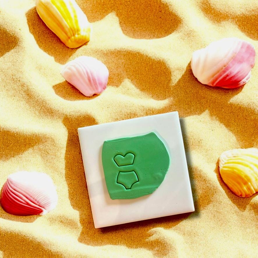 Polymer clay cutouts of bikini top and bottom displayed on a white ceramic tile with beach decor.