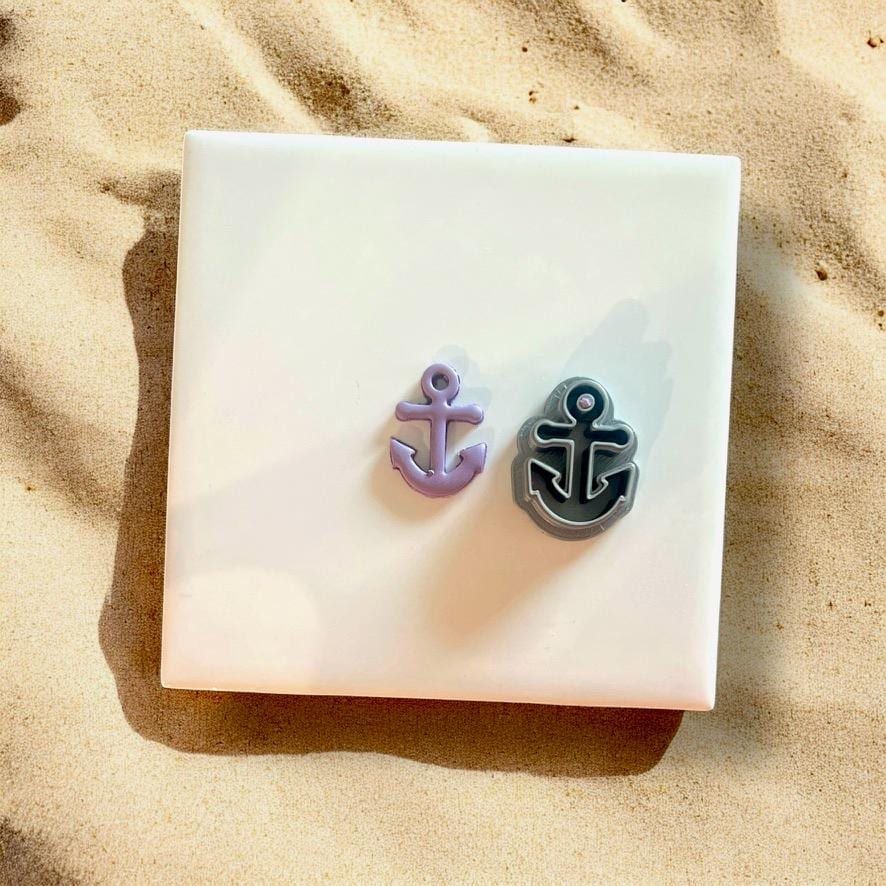 Two anchor-shaped clay cutters in purple and gray, alongside their clay impressions on a white tile with beach sand..