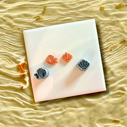 Polymer clay fish and shell charms alongside their cutters in gray, displayed on a white tile with coastal water backdrop.