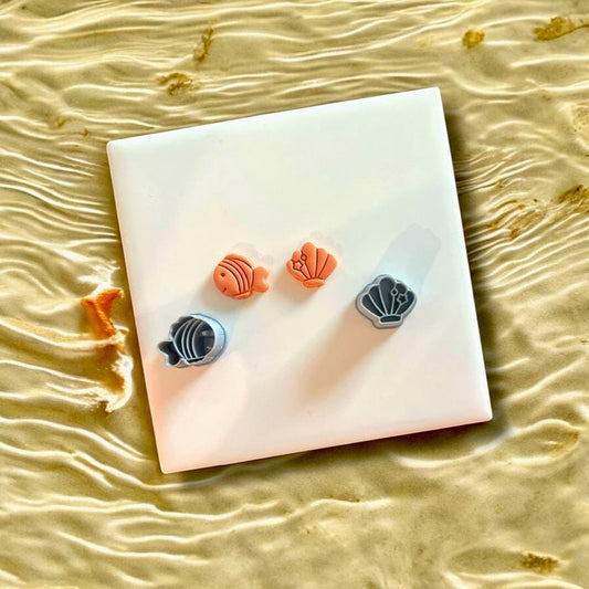 Polymer clay fish and shell charms alongside their cutters in gray, displayed on a white tile with coastal water backdrop.