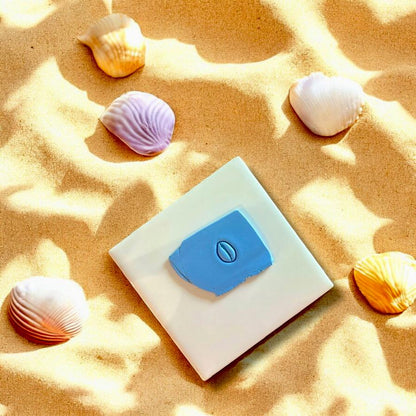 Cowrie shell clay imprint on blue polymer clay, displayed on white tile with seashells and beach sand accents.