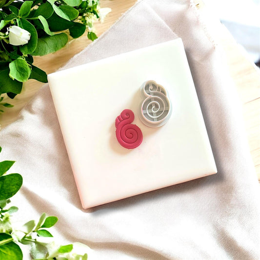 Flamingo clay charm alongside its matching cutter, displayed on a white tile with fresh greenery and a fabric backdrop.