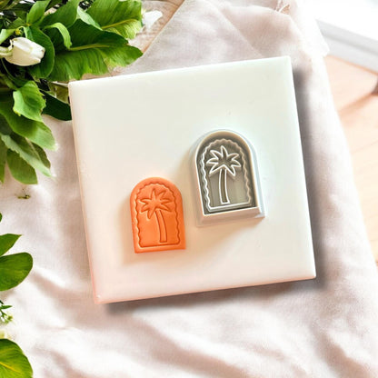 Tropical palm tree arch clay charms and their matching cutter, displayed on a white tile with a soft fabric backdrop.