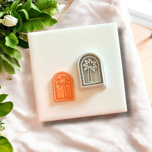 Tropical palm tree arch clay charms and their matching cutter, displayed on a white tile with a soft fabric backdrop.