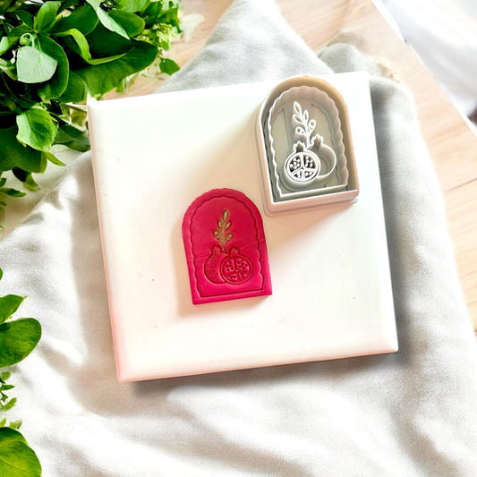 Polymer clay pomegranate arch charm alongside its matching cutter, displayed on a white tile with fresh greenery.