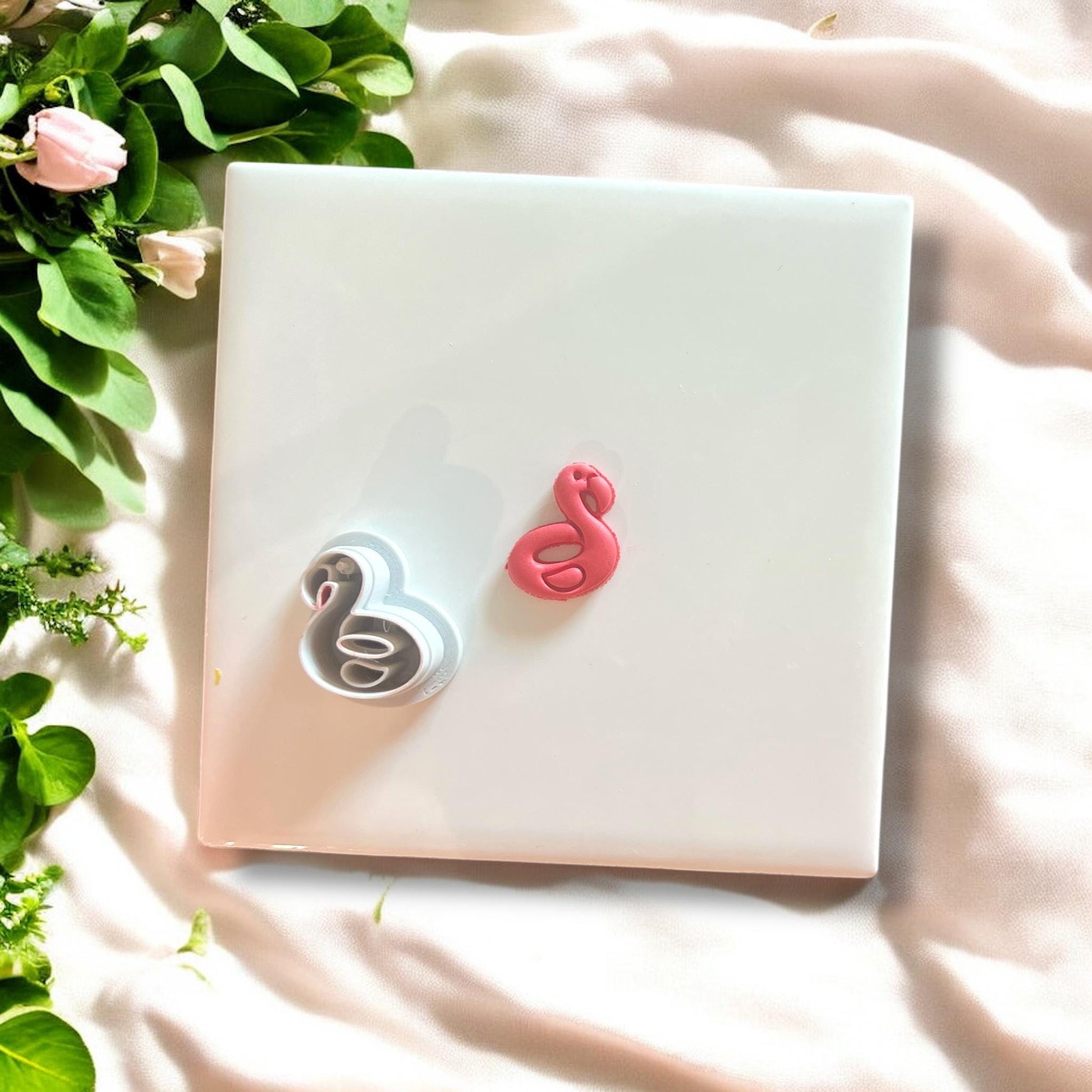 Side-by-side view of the flamingo floatie polymer clay cutter and pink clay charm on a white tile with leaves.