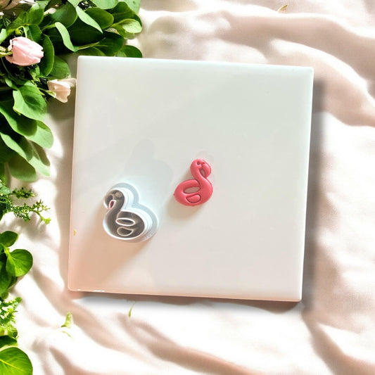Side-by-side view of the flamingo floatie polymer clay cutter and pink clay charm on a white tile with leaves.