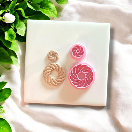 Swirl shell polymer clay charms alongside pink shell-shaped cutters, displayed on white tile.