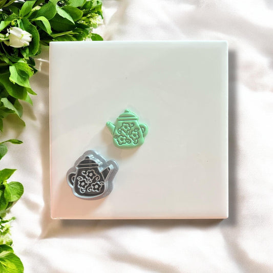 Floral teapot charm next to the matching cutter, displayed on a white tile with greenery.