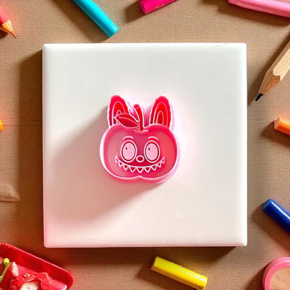 Pink apple monster clay cutter centered on white tile with pencils