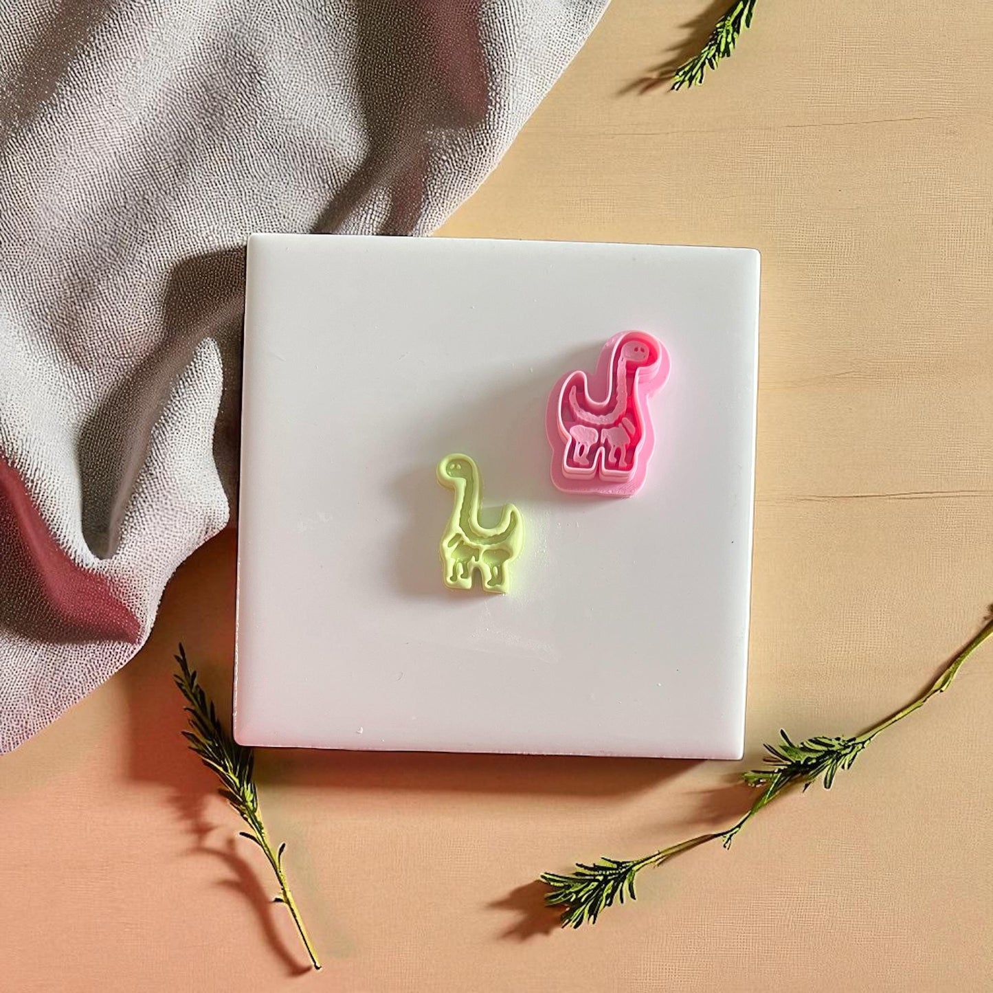 Pink dinosaur cutter next to clay imprint on tile with fabric and greens