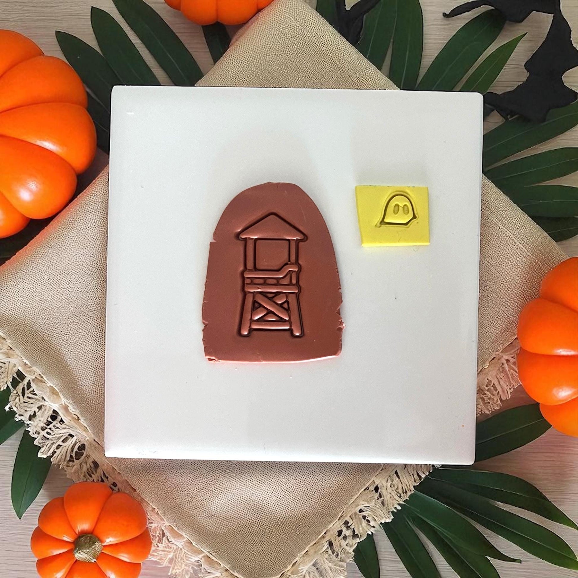 Brown clay impression of lifeguard tower with yellow ghost shape on white tile and pumpkins.