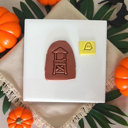 Brown clay impression of lifeguard tower with yellow ghost shape on white tile and pumpkins.