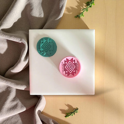 Pink and teal round clay cutters with tribal diamond designs arranged on white tile with fabric backdrop.