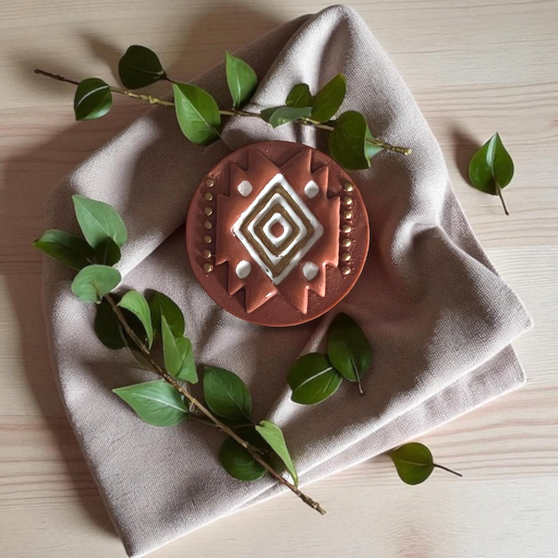 Round terracotta-colored polymer clay cutter with tribal diamond design placed on beige fabric and green leaves.