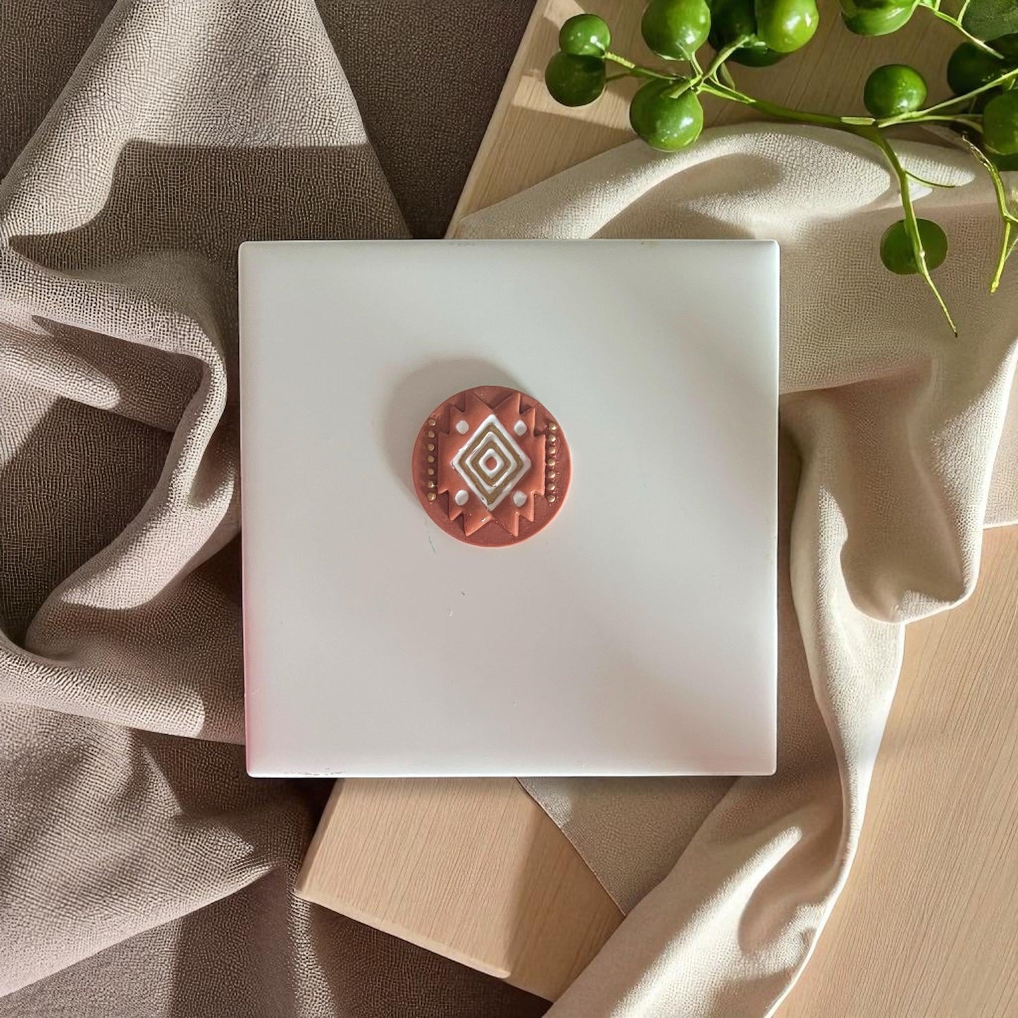 Round terracotta-colored polymer clay cutter with tribal diamond design placed on beige fabric and green leaves.