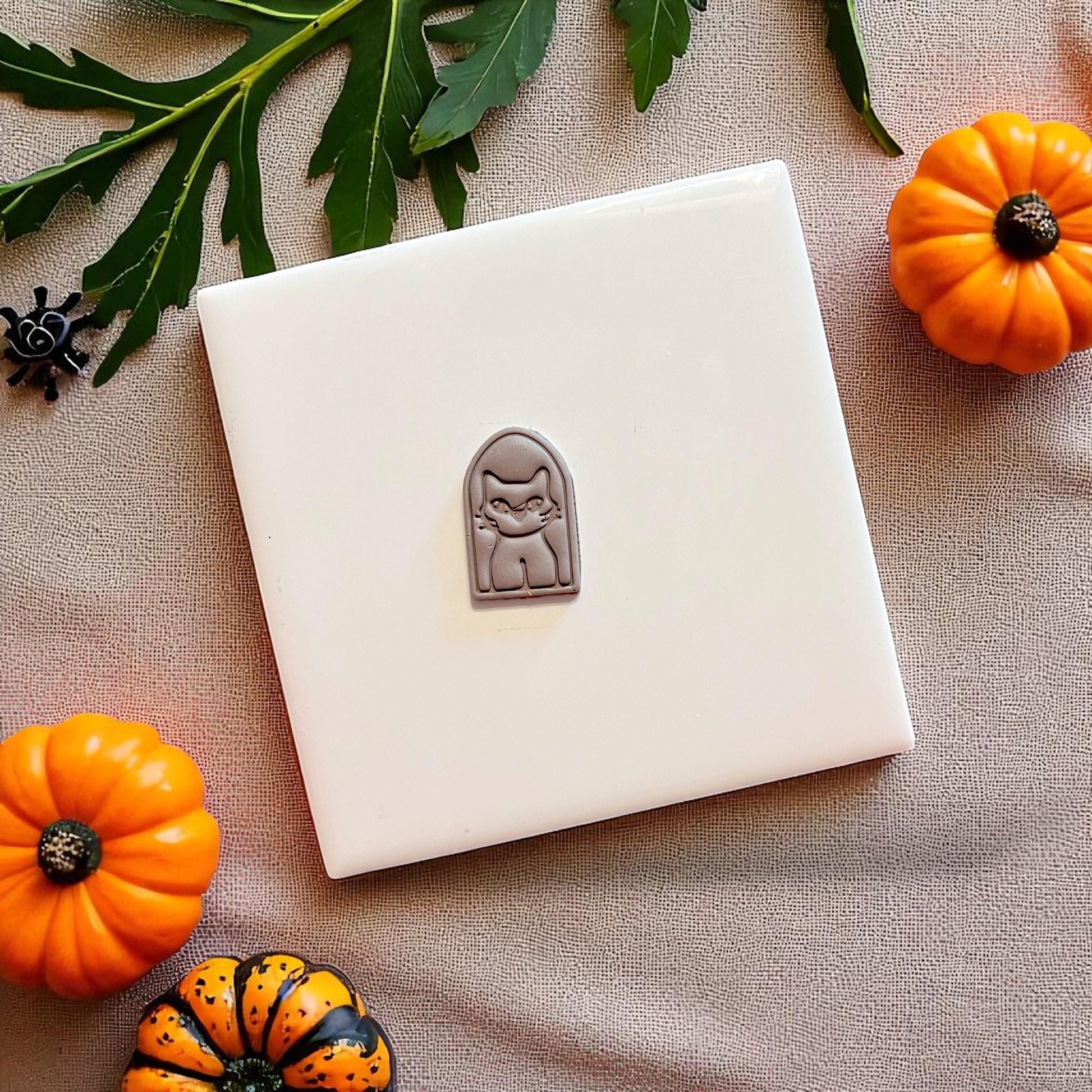 Small clay cutout of spooky cat portrait on white tile surrounded by pumpkins