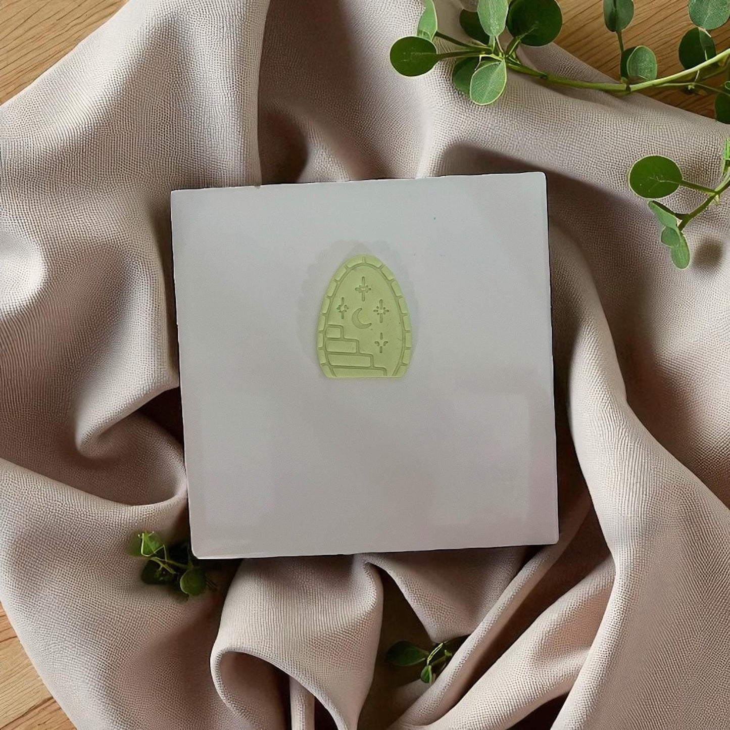 Small green arched clay imprint with stairway, moon, and stars on white tile with beige fabric.