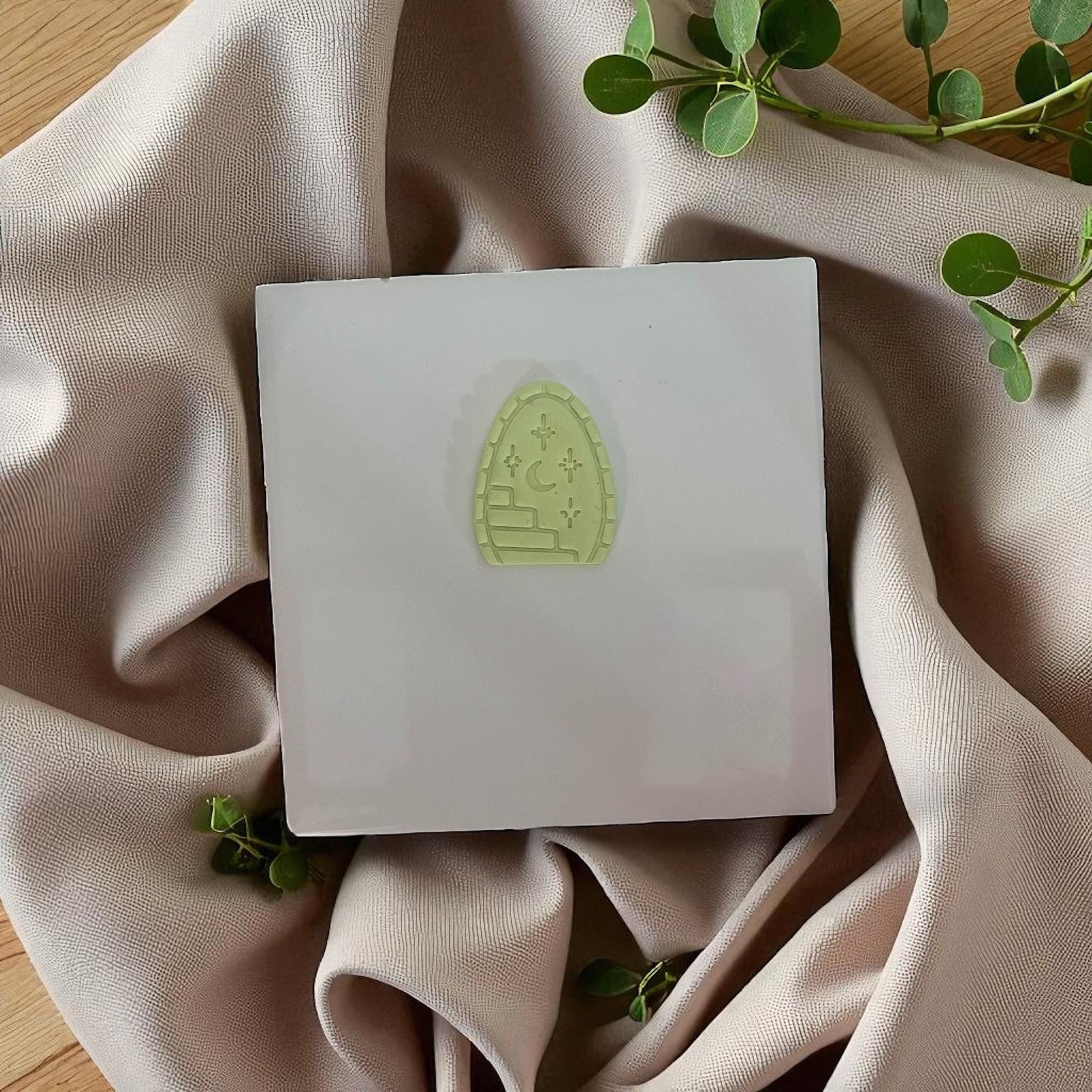 Small green arched clay imprint with stairway, moon, and stars on white tile with beige fabric.