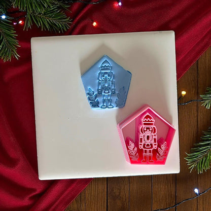 Blue nutcracker clay imprint and pink nutcracker cutter side by side on white tile with Christmas background.