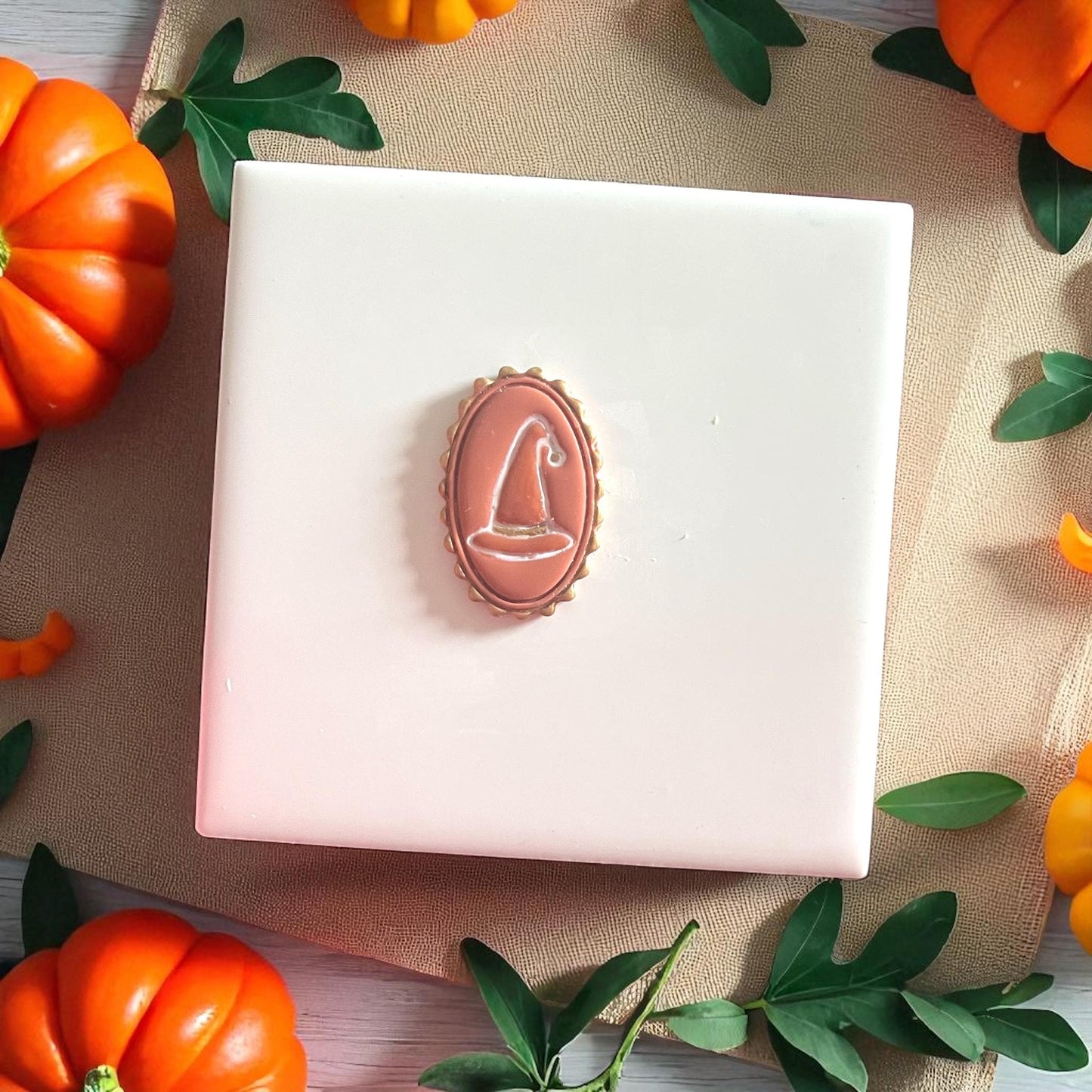 Brown witch hat clay imprint in oval cameo frame on white tile with pumpkins and greenery.