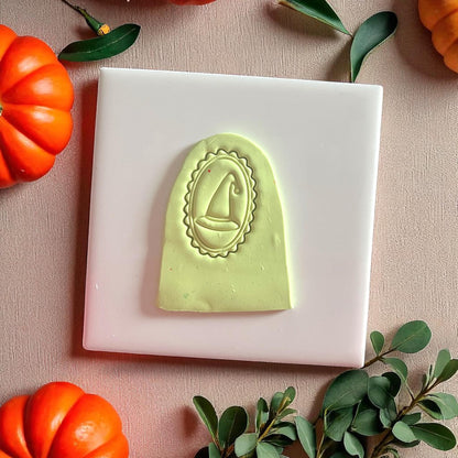 Green clay imprint of oval cameo witch hat on white tile surrounded by pumpkins and leaves.