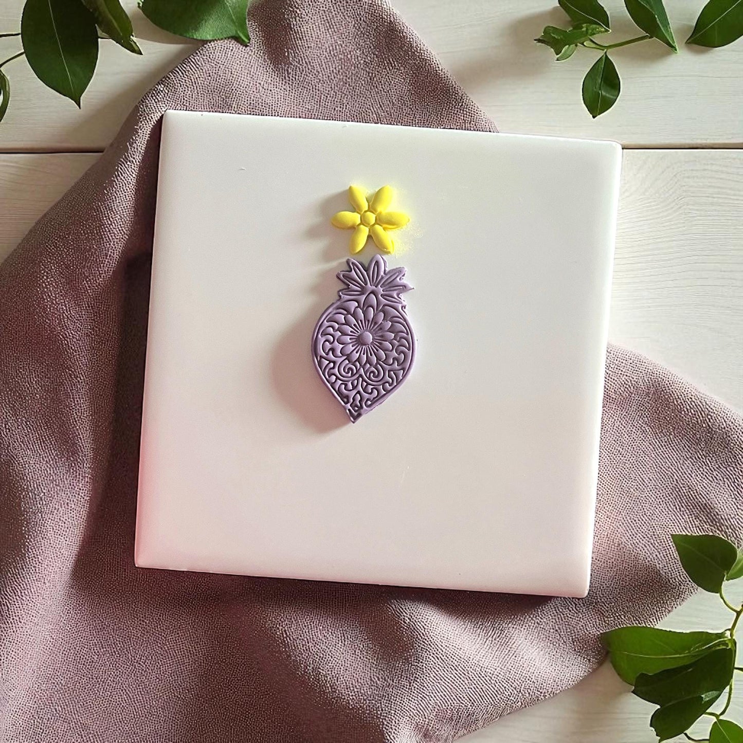 Purple mandala flower clay imprint with yellow flower imprint on white tile and greenery.