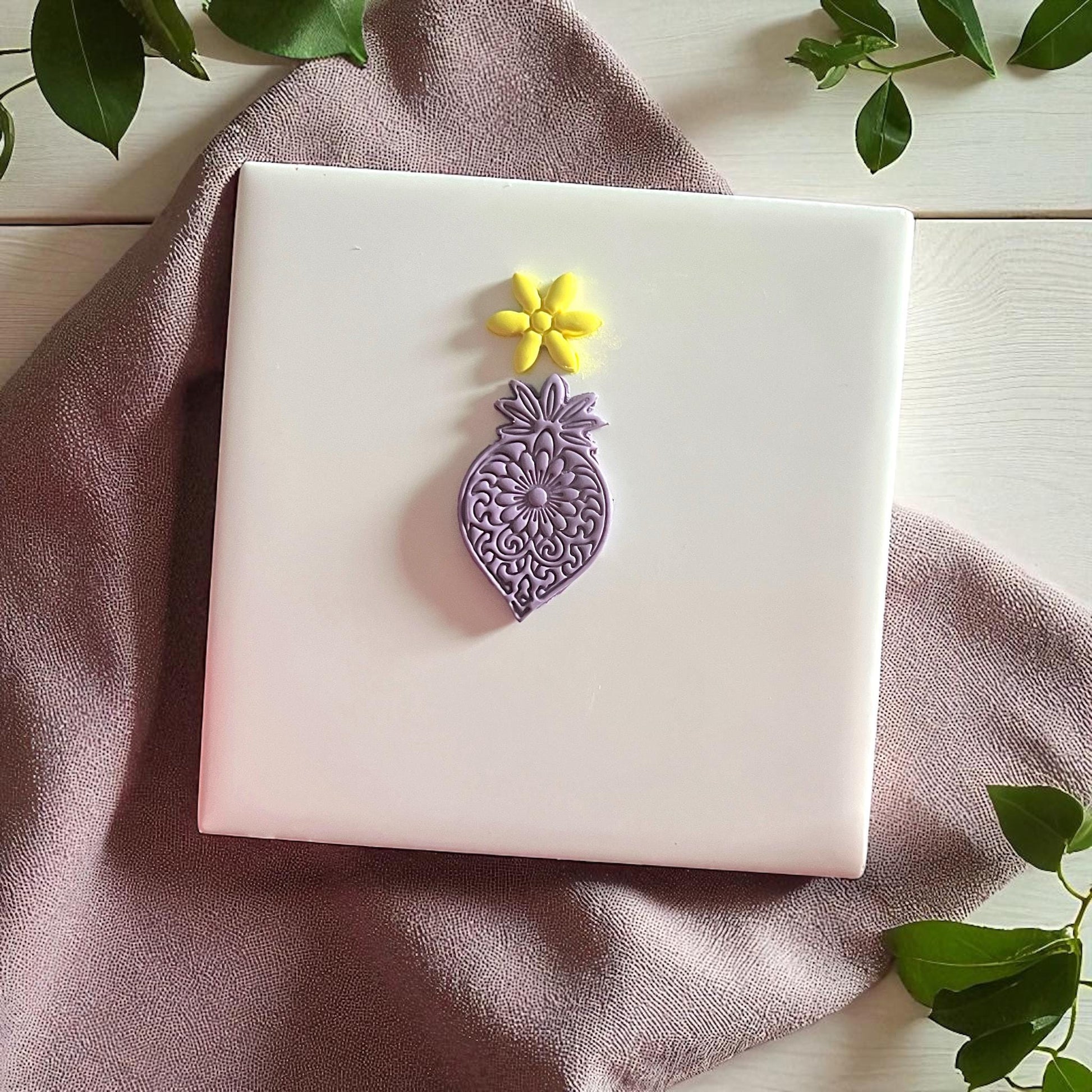 Purple mandala flower clay imprint with yellow flower imprint on white tile and greenery.