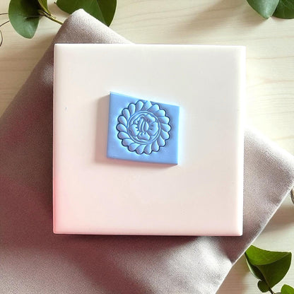 Blue clay imprint of round flower design on white tile with beige fabric and leaves.