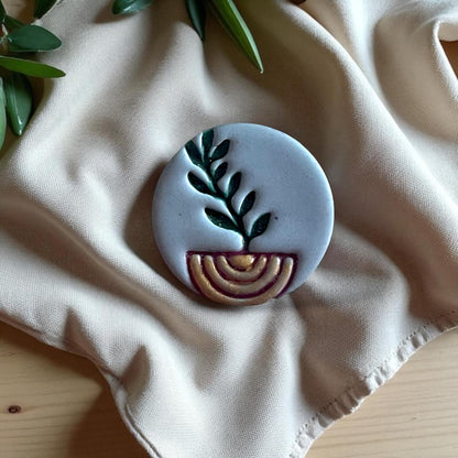 Large painted clay piece with green plant stem and golden arch on beige fabric.