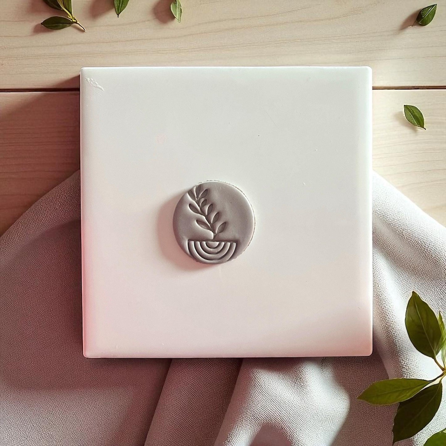 Round gray clay imprint with plant and arch design on white tile and beige fabric.