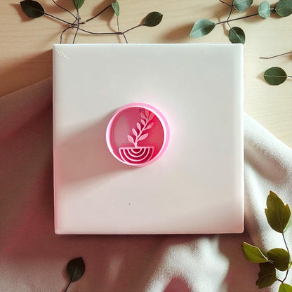 Pink plant arch clay cutter on white tile with greenery and fabric background.