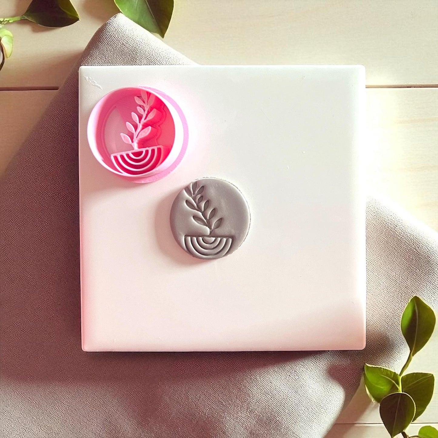 Pink plant arch cutter next to gray clay imprint on white tile with fabric and greenery.