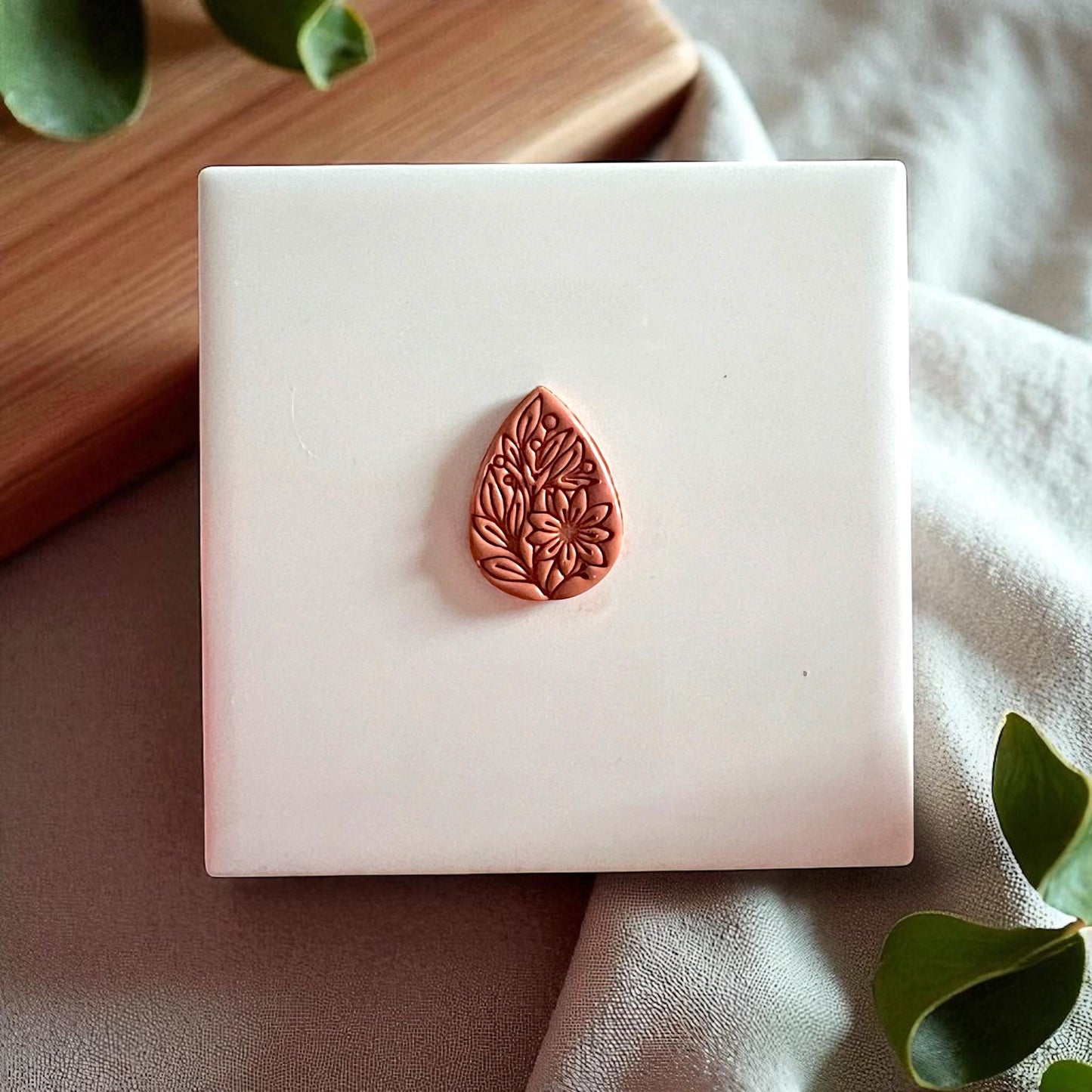 Brown floral teardrop clay imprint centered on white tile with fabric and wood background.
