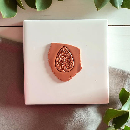 Brown clay imprint of floral teardrop design on white tile with fabric and greenery.