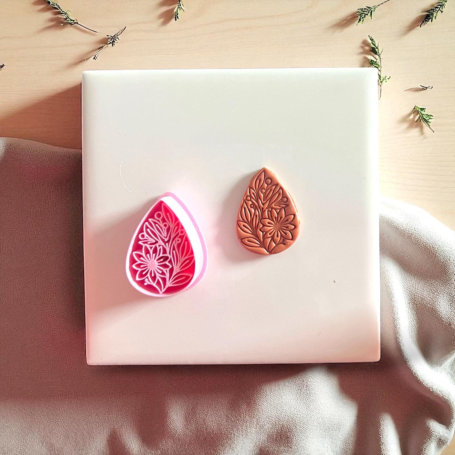 Pink floral teardrop cutter beside brown clay imprint on white tile with fabric background.