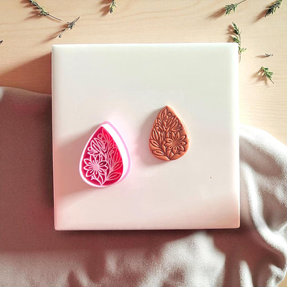 Pink floral teardrop cutter beside brown clay imprint on white tile with fabric background.