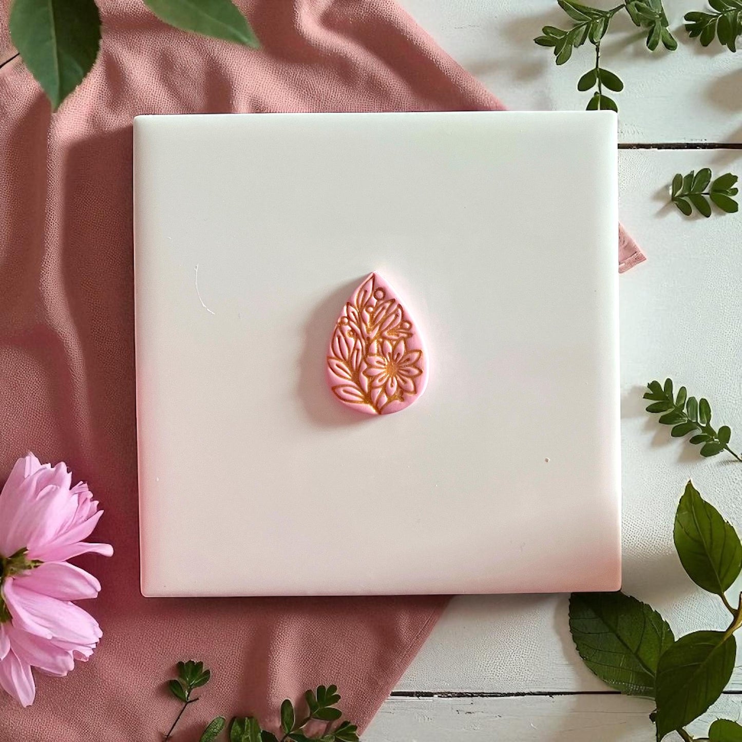 Pink floral teardrop clay imprint with gold painted details on white tile with greenery.