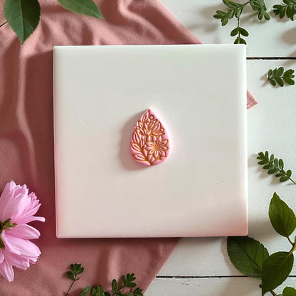 Pink floral teardrop clay imprint with gold painted details on white tile with greenery.