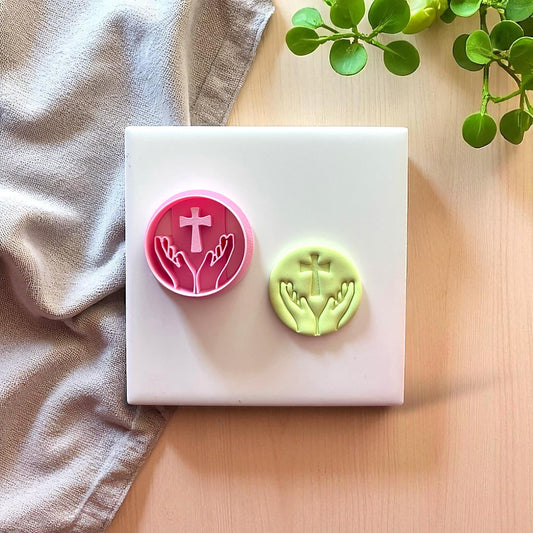 Pink cutter and green clay imprint of hands with cross on white tile with fabric and plant.