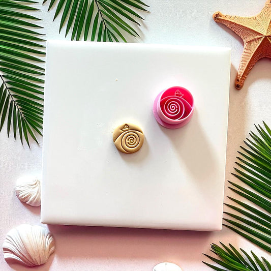 Pink paper boat cutter beside tan clay imprint with spiral wave design on white tile.