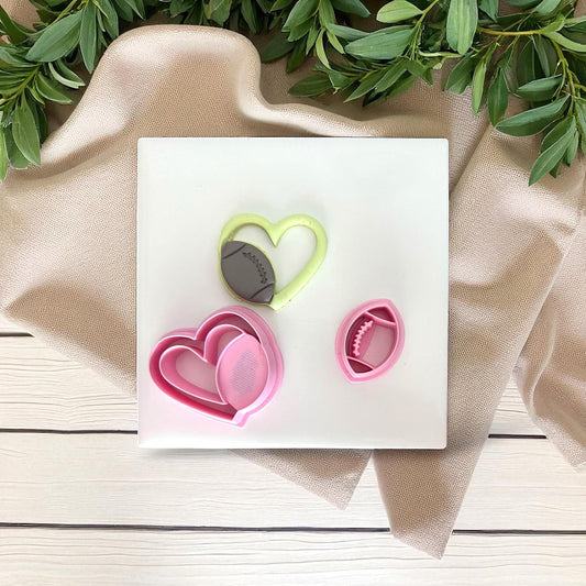 Green football heart clay piece beside pink cutter set on white tile with fabric and greenery.