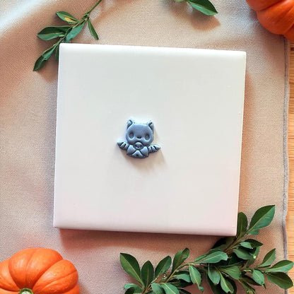 Small gray baby bat clay piece on white tile with pumpkins and greenery.