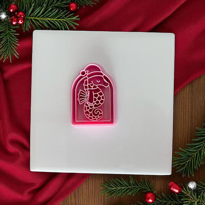 Pink Christmas seahorse clay cutter on white tile with red fabric and pine branches.