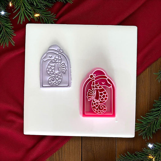 Purple seahorse clay imprint beside pink cutter on white tile with holiday background.