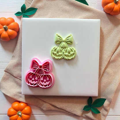 Pink pumpkin cherry bow cutter with green imprint on white tile and pumpkins.