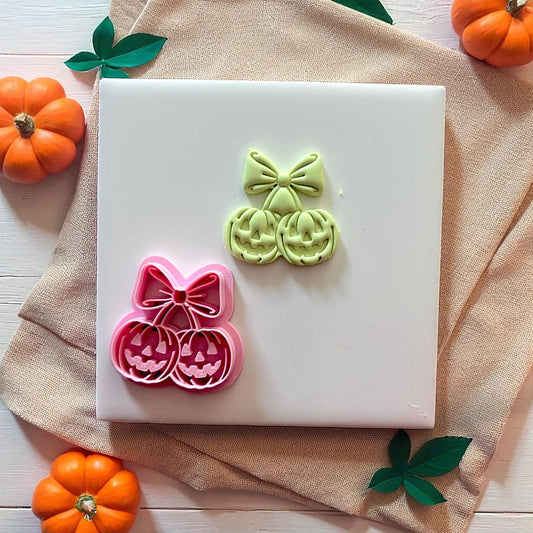 Pink pumpkin cherry bow cutter with green imprint on white tile and pumpkins.