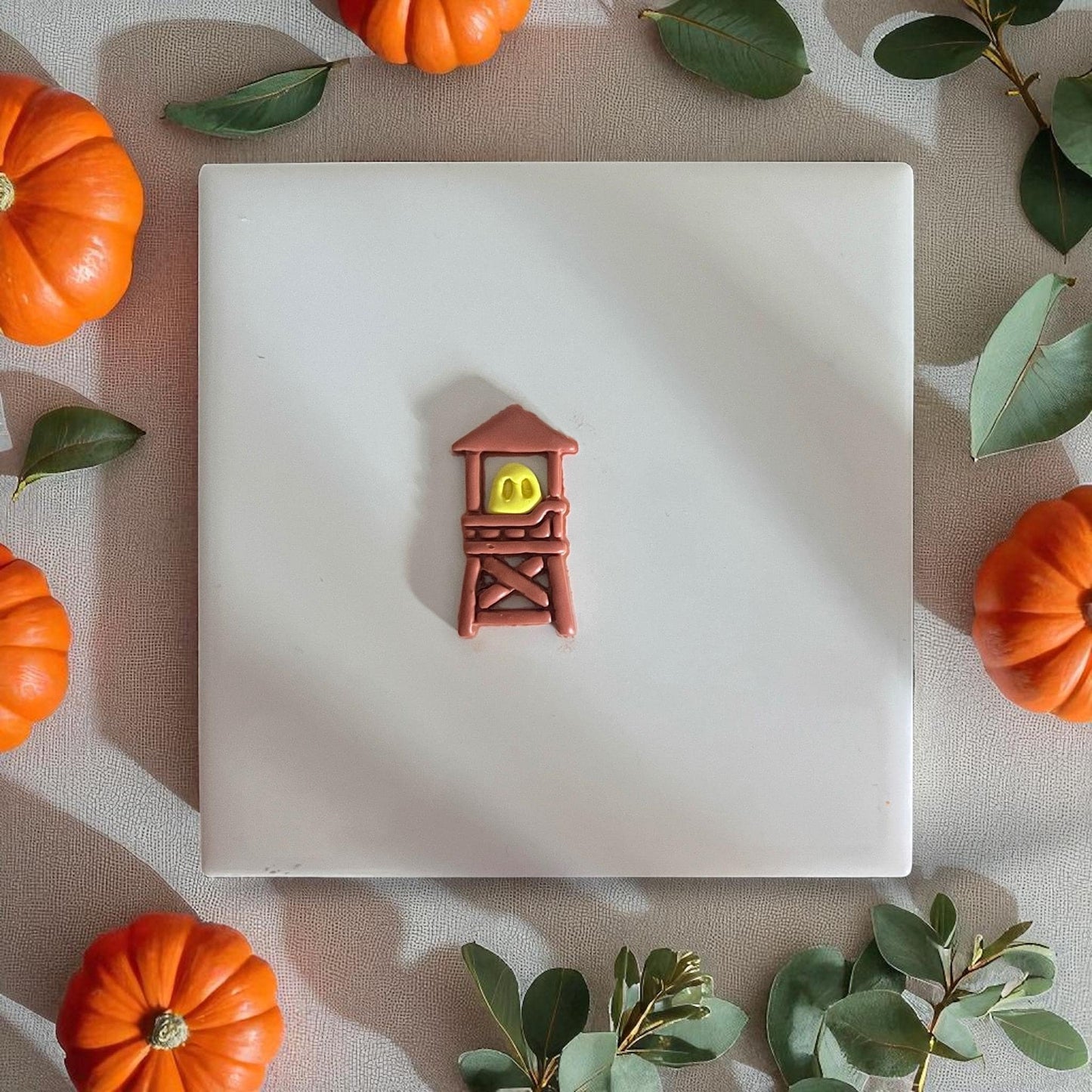Small ghost inside brown clay tower displayed on white tile with pumpkins and leaves.