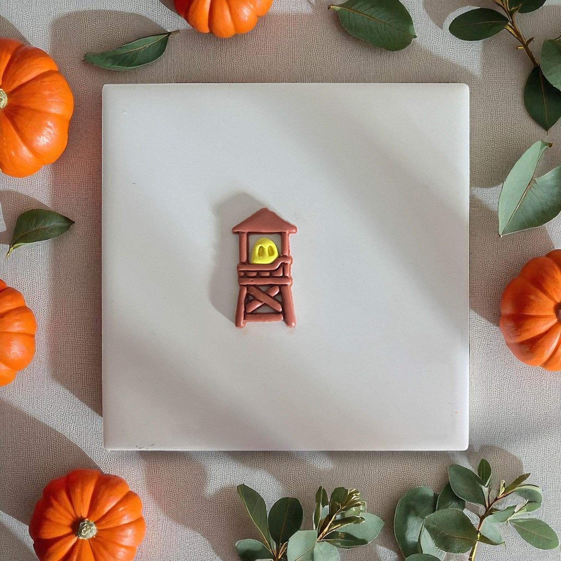 Small ghost inside brown clay tower displayed on white tile with pumpkins and leaves.