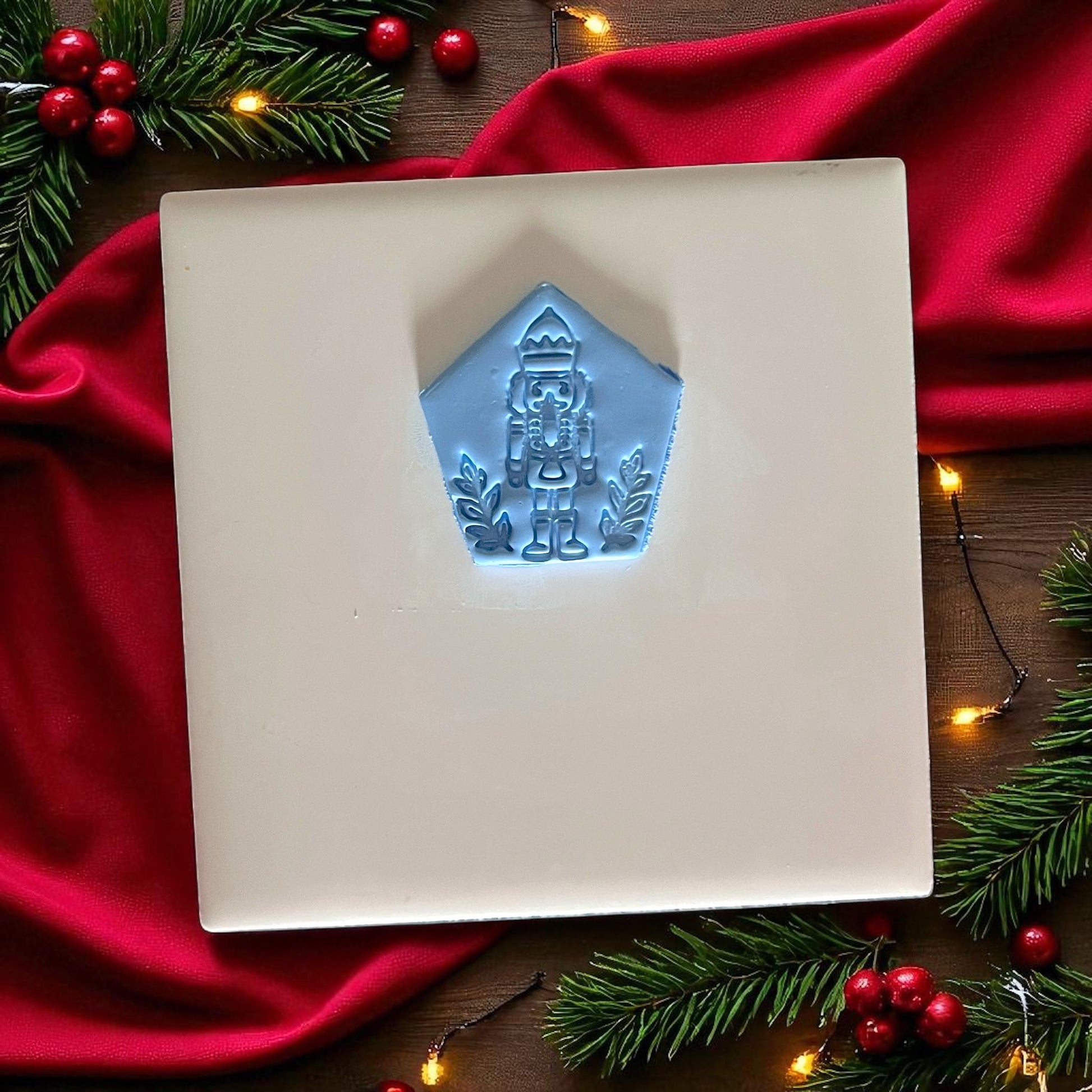 Blue nutcracker clay imprint on white tile with red fabric, pine branches, and festive lights.
